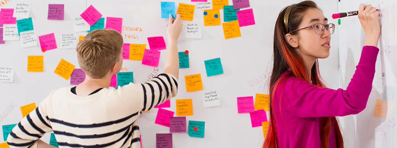 Photo of two students writing on post-it notes on a whiteboard