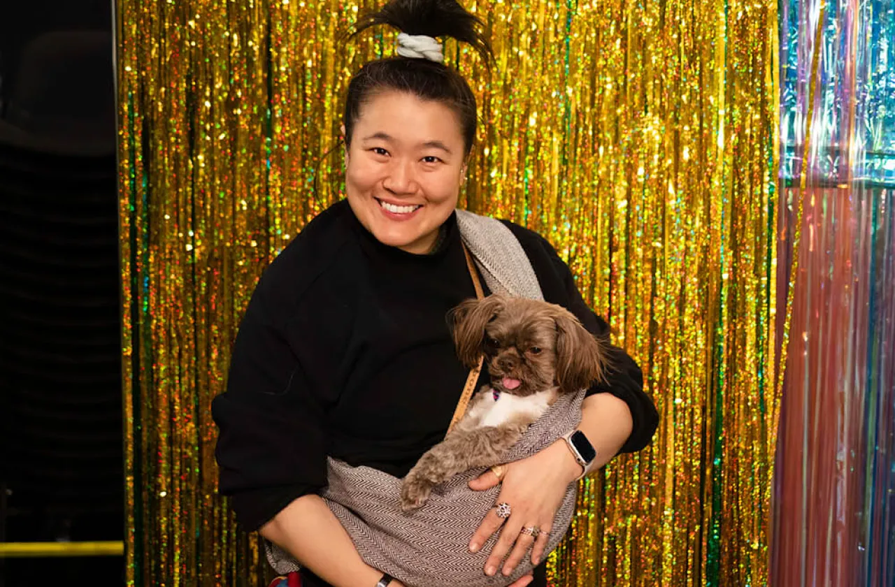 Theater artist Diana Oh holds their dog shovels against a backdrop of shimmering beaded curtains