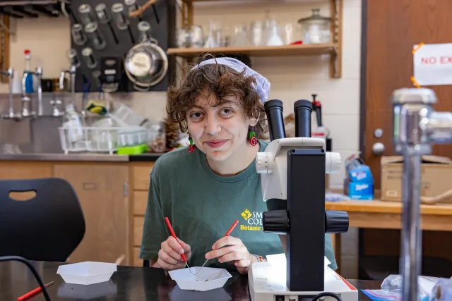 student with brown hair, purple bandana and green shirt in front of a microscope