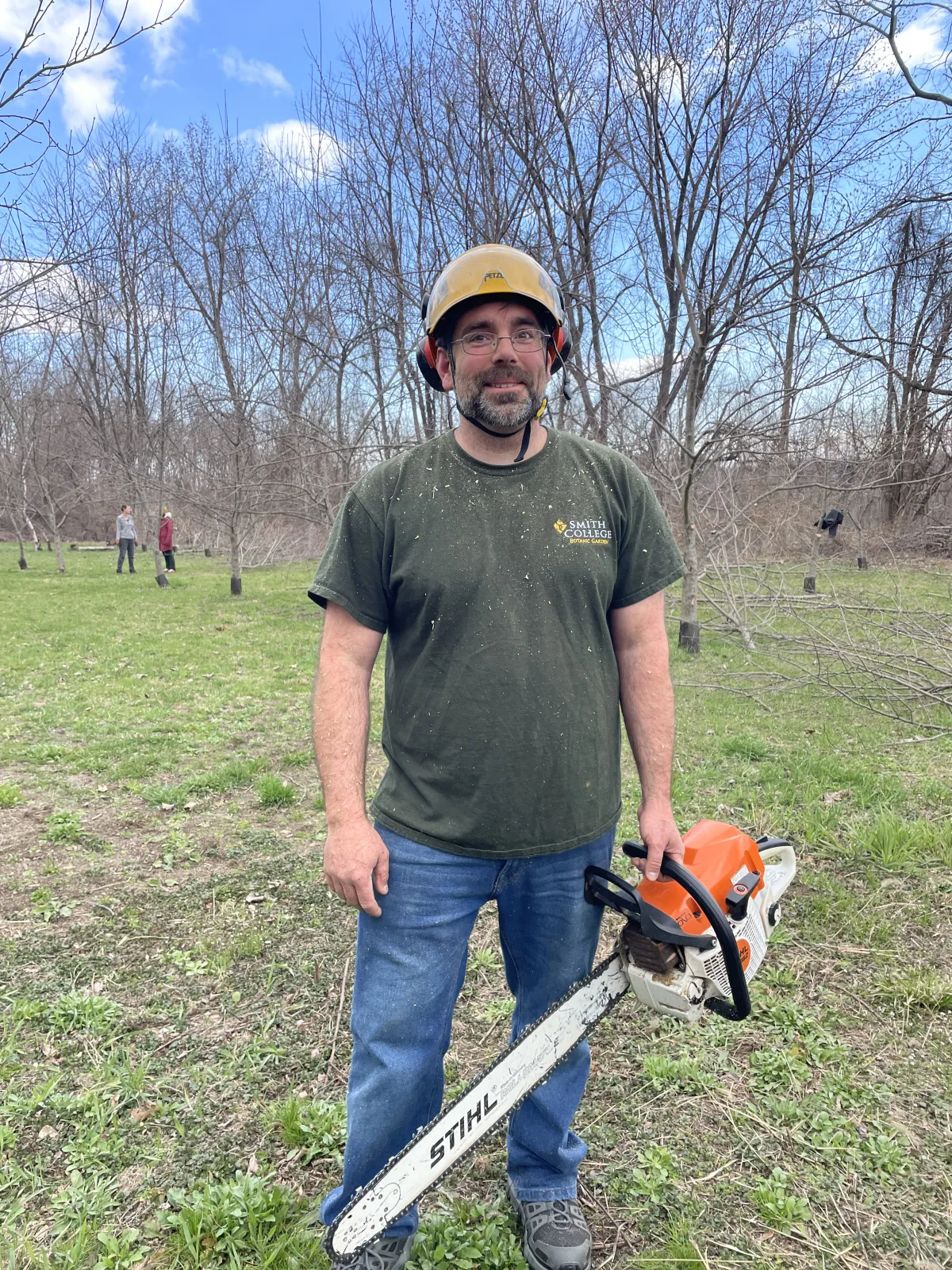 Man with yellow safety hat and chainsaw in green shirt.