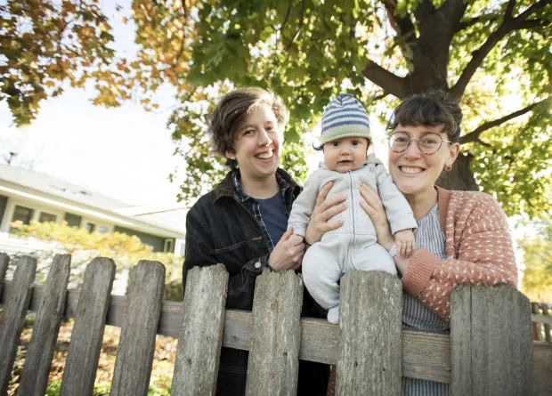 A photo of two smiling alums holding their baby daughter between them, smiling near a fence on a sunny day with a tree behind them.