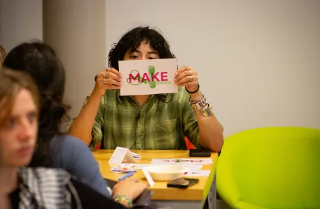 A student holds a paper with the words Make in front of their face while looking at the camera at ZineFest 2024