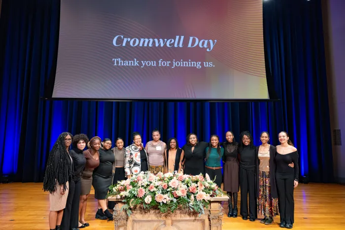 Several students pose for a photo while standing on the stage in front of a large screen that reads, "Cromwell Day. Thank you for joining us." President Sarah Willie-LeBreton poses with them in the center.