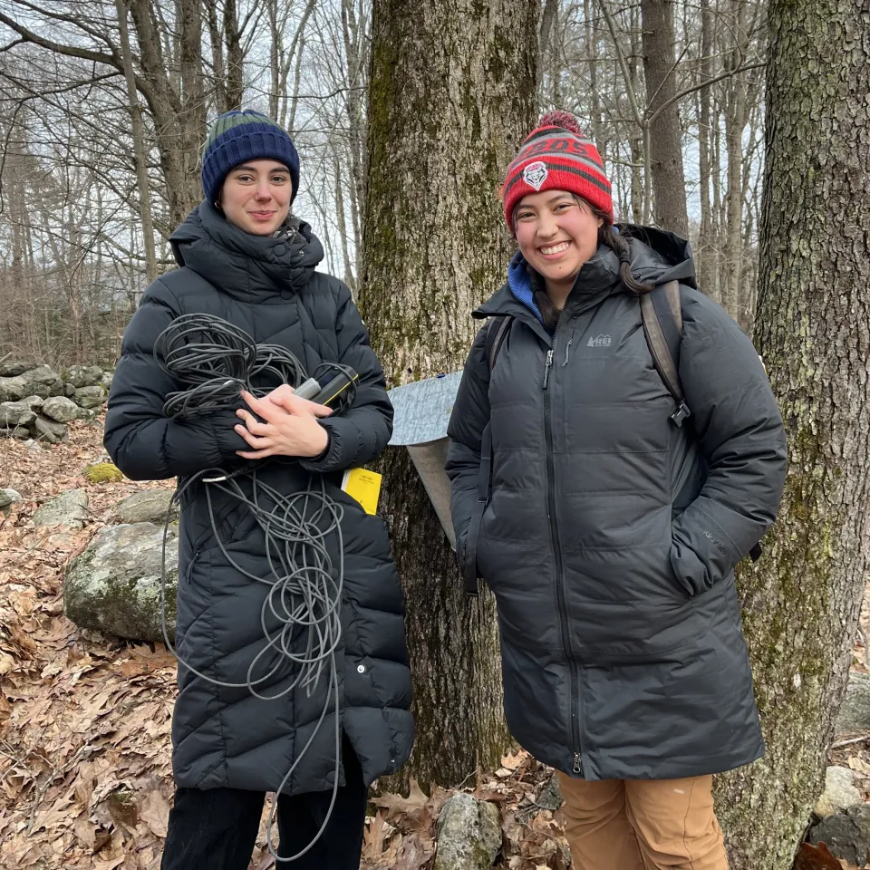 Two smiling students standing in winter clothes in front of trees, surrounded by fallen leaves, holding equipment