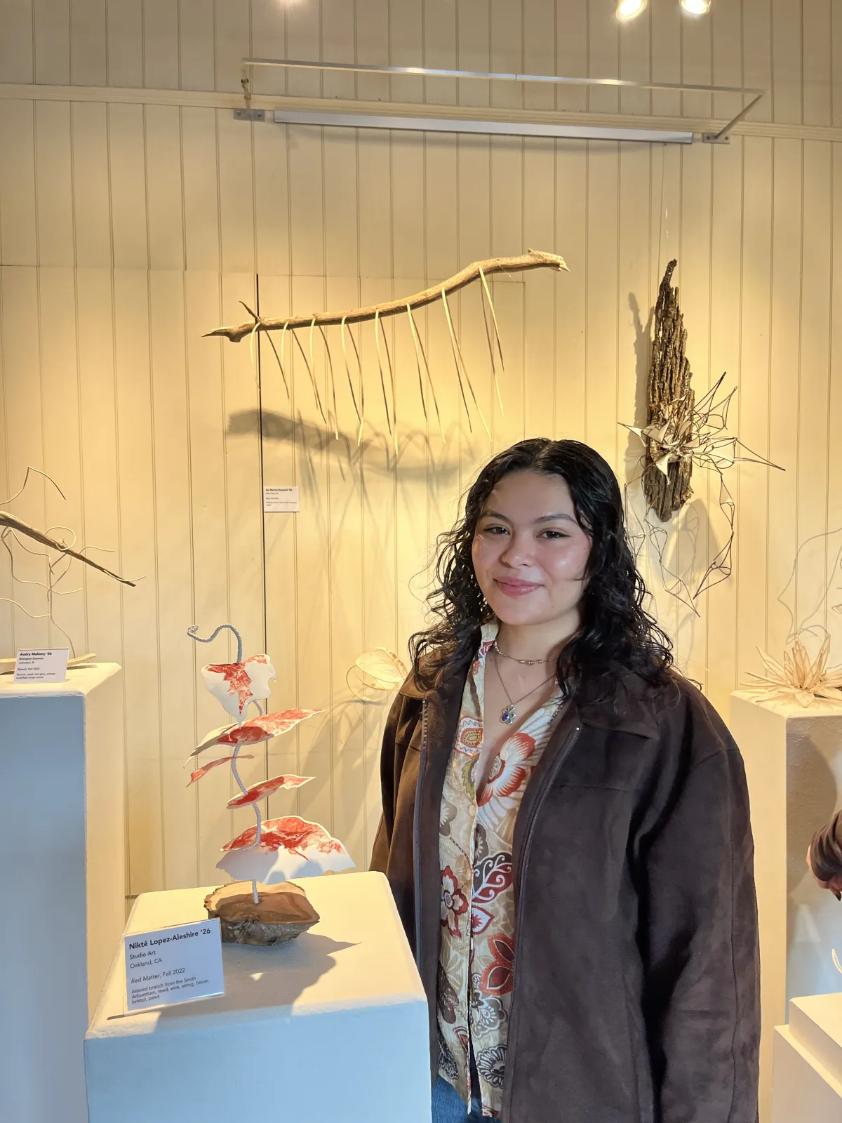 woman with brown curly hair and brown leather jacket posing in front of small scultpure.